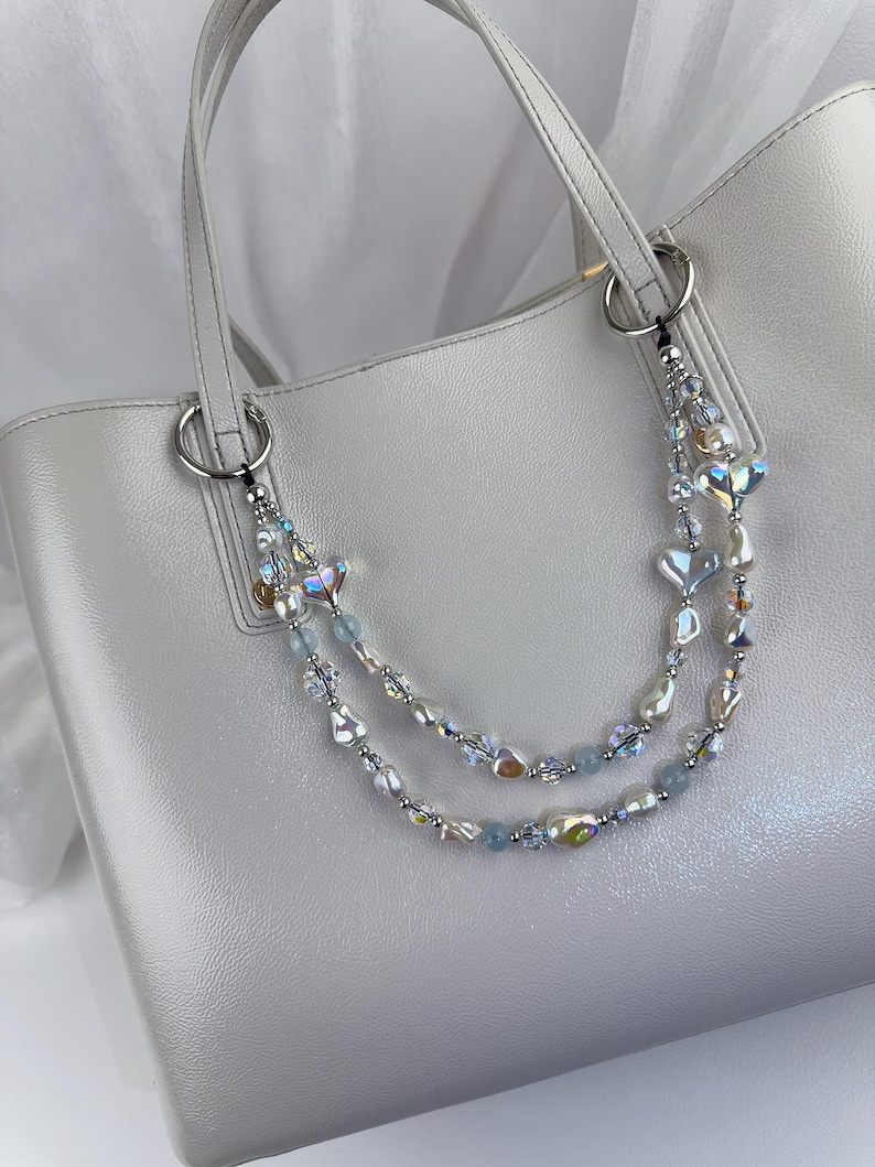 May include: A light gray tote bag featuring a decorative double-strand beaded chain attached to the silver-toned rings. The chain consists of iridescent heart-shaped beads, white faux pearls, small clear crystals, and light blue round beads.
