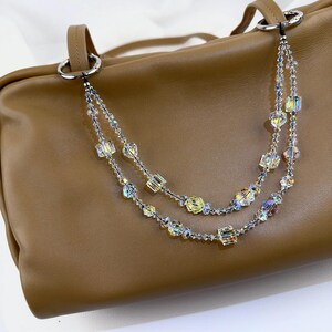 May include: A tan leather handbag featuring a decorative double-strand beaded chain accessory. The chain is composed of clear, iridescent, and yellow faceted crystal beads in various shapes, attached to the bag's silver-toned metal rings.