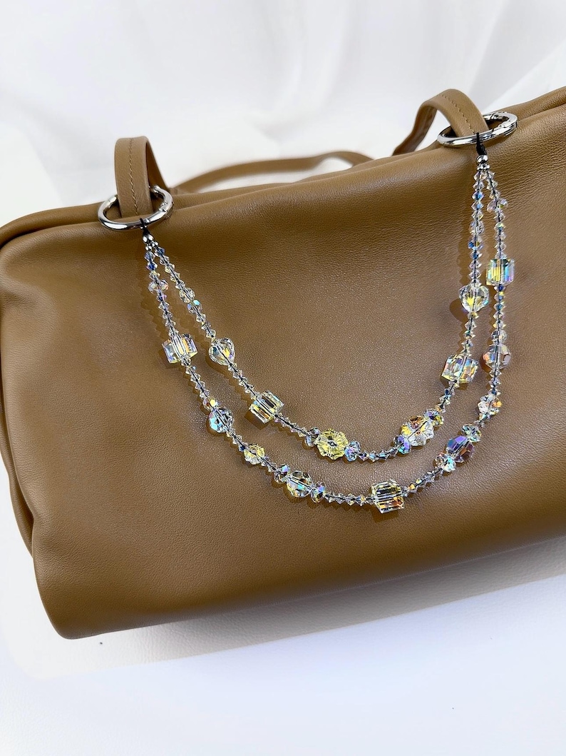 May include: A tan leather handbag featuring a decorative double-strand beaded chain accessory. The chain is composed of clear, iridescent, and yellow faceted crystal beads in various shapes, attached to the bag's silver-toned metal rings.