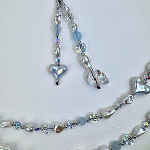May include: A beaded accessory featuring a silver-toned metal ring attached to two dangling strands of iridescent heart-shaped beads, clear faceted crystals, light blue round beads, and irregular white pearl-like beads with a metallic finish.