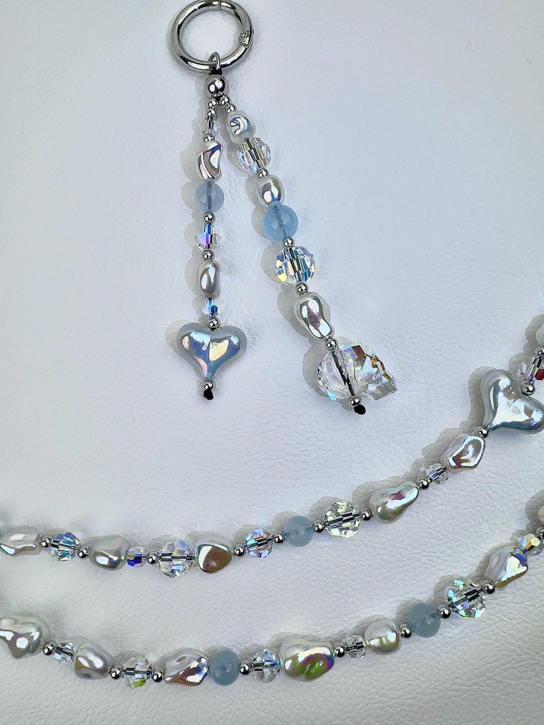 May include: A beaded accessory featuring a silver-toned metal ring attached to two dangling strands of iridescent heart-shaped beads, clear faceted crystals, light blue round beads, and irregular white pearl-like beads with a metallic finish.