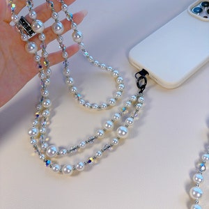 May include: A white pearl and crystal phone strap with a silver clasp. The strap is made of multiple strands of pearls and crystals.