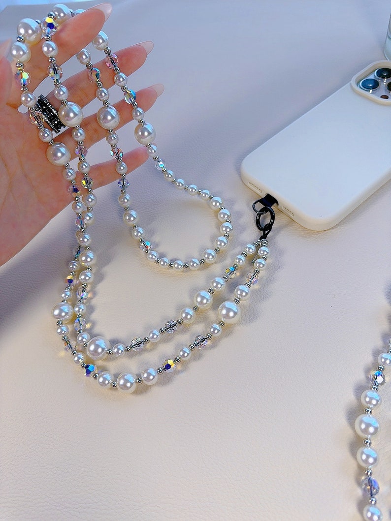 May include: A white pearl and crystal phone strap with a silver clasp. The strap is made of multiple strands of pearls and crystals.
