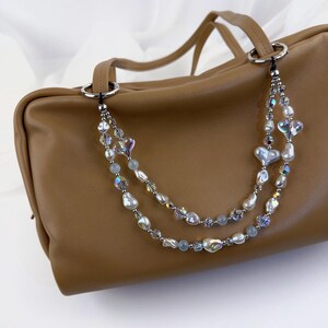 May include: A tan leather handbag featuring a decorative double-strand beaded chain strap. The strap is adorned with iridescent heart-shaped beads, white baroque pearls, small clear faceted crystals, and light blue round beads attached to silver rings.