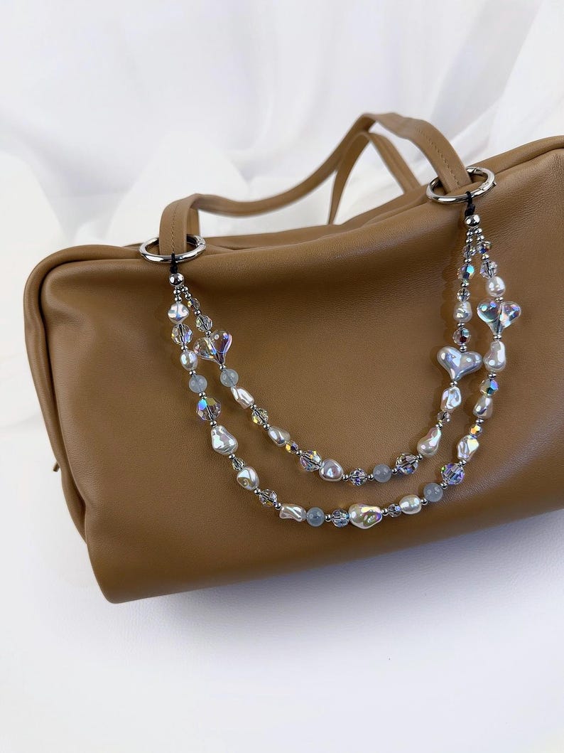 May include: A tan leather handbag featuring a decorative double-strand beaded chain strap. The strap is adorned with iridescent heart-shaped beads, white baroque pearls, small clear faceted crystals, and light blue round beads attached to silver rings.