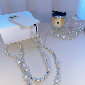 May include: A white phone case with a pearl and crystal phone strap. The strap is a double strand with large white pearls and smaller clear crystals.