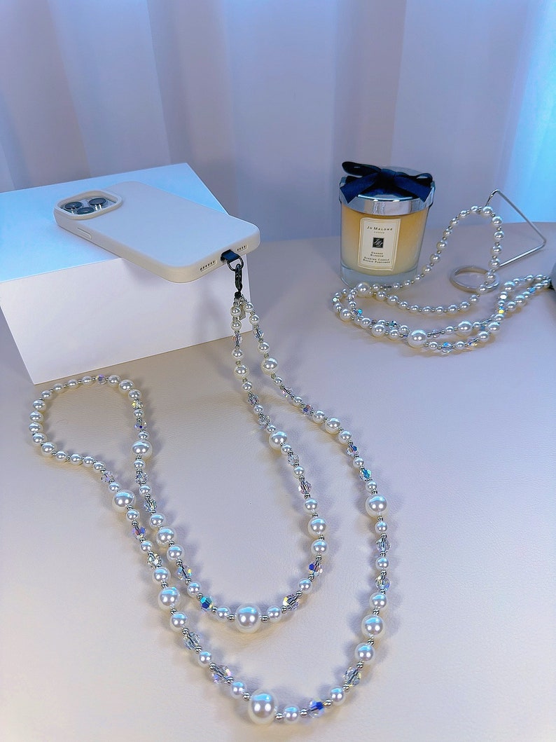 May include: A white phone case with a pearl and crystal phone strap. The strap is a double strand with large white pearls and smaller clear crystals.