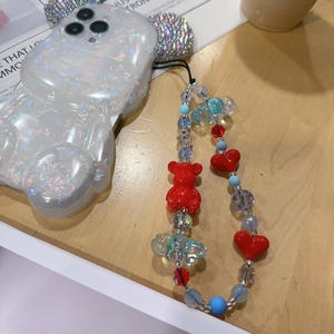 May include: A white iridescent phone case shaped like a bear with a clear phone strap attached. The strap has red heart charms, red bear charms, and clear and blue beads.