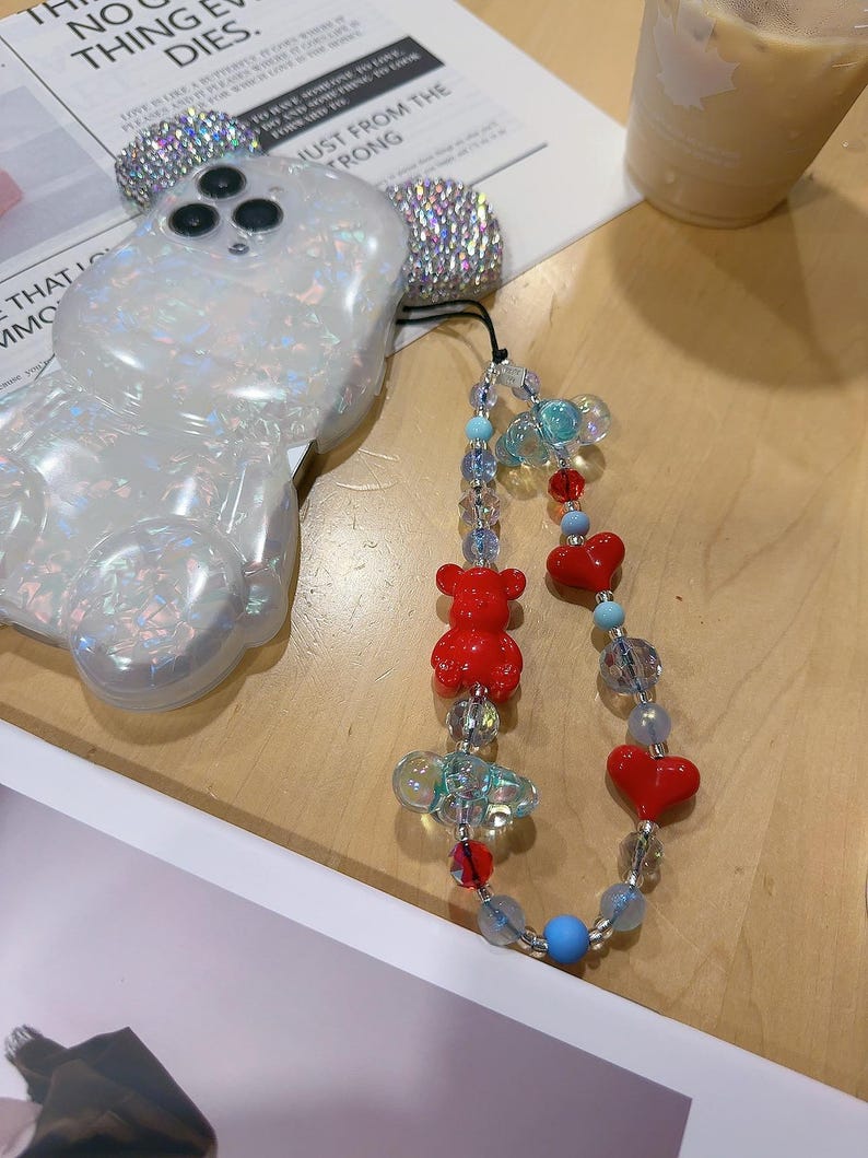 May include: A white iridescent phone case shaped like a bear with a clear phone strap attached. The strap has red heart charms, red bear charms, and clear and blue beads.