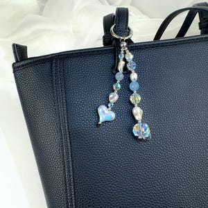 May include: A black textured handbag featuring a decorative charm attached to the handle. The charm consists of two strands of beads, including iridescent heart-shaped beads, clear faceted crystals, and smooth light blue spherical beads.