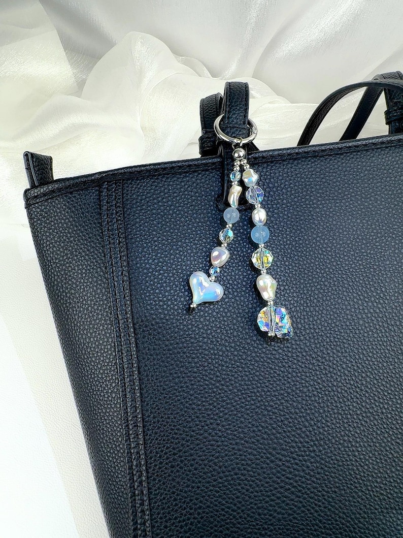 May include: A black textured handbag featuring a decorative charm attached to the handle. The charm consists of two strands of beads, including iridescent heart-shaped beads, clear faceted crystals, and smooth light blue spherical beads.