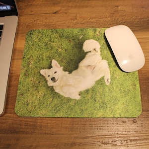 May include: A rectangular mousepad featuring a white dog lying on a green grass background. A white computer mouse is next to the mousepad. A laptop is partially visible on a wooden desk.