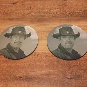 May include: Two round coasters featuring a black and white image of a man in a cowboy hat. The coasters are made of a dark material and are placed on a wooden surface.