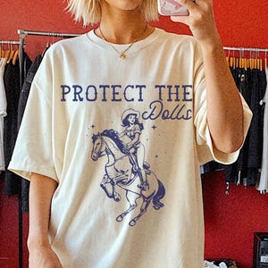 Puede incluir: Camiseta crema de gran tamaño con el texto "PROTECT THE Dolls" y un gráfico azul de una vaquera montando a caballo. La modelo lleva botas de vaquero rosas. El fondo incluye percheros y una pared roja.