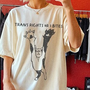 May include: Cream-colored t-shirt with the text "TRANS RIGHTS OR I BITES" and a black and white graphic of a dog. The shirt is oversized and worn with pink boots. Clothing items hang on racks in the background.