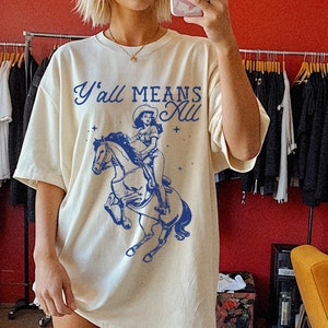 以下が含まれることがあります： 「Y'all Means All」の文字と、カウガールが馬に乗っている青いグラフィックがプリントされた、クリーム色のオーバーサイズのTシャツ。明るいピンクのカウボーイブーツがコーディネートされています。カジュアルで快適なスタイルです。