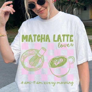 May include: White t-shirt with a green graphic of a matcha latte and a whisk. The text on the shirt reads "Matcha Latte lover 6 am-11 am every morning".