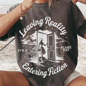 K&ouml;nnte beinhalten: Dunkelgraues T-Shirt mit dem Text "Leaving Reality Entering Fiction" und einer Grafik einer Frau an einer T&uuml;r. Das Shirt hat kurze &Auml;rmel und einen Rundhalsausschnitt. Der Text "It's a Flare Day" steht ebenfalls auf dem Shirt.