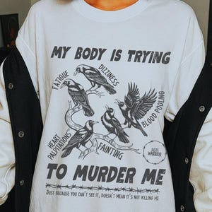以下が含まれることがあります： 「MY BODY IS TRYING TO MURDER ME」の文字と、疲労やめまいなどの症状を示す鳥のグラフィックが描かれた白いTシャツ。白と黒のジャケットと合わせて着用されています。