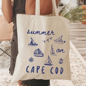 Puede incluir: Un bolso tote de color crema con ilustraciones náuticas azules y las palabras "summer on Cape Cod". El bolso tiene asas largas y está hecho de una tela resistente. El diseño incluye veleros, un ancla y otros gráficos temáticos de verano.