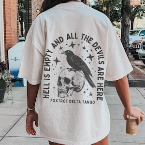 Puede incluir: Camiseta crema de gran tamaño con un gráfico en la espalda que presenta un cuervo posado sobre una calavera, rodeado de estrellas y el texto "HELL IS EMPTY AND ALL THE DEVILS ARE HERE". La camiseta se usa al aire libre.