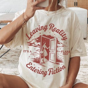 K&ouml;nnte beinhalten: Cremefarbenes T-Shirt mit dem Text "Leaving Reality Entering Fiction" in Rot. Die Grafik zeigt eine Person, die durch eine T&uuml;r geht. Das Shirt wird mit wei&szlig;en Shorts und schwarz-wei&szlig; gestreiften Socken getragen.