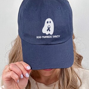 May include: Navy blue baseball cap with a white ghost graphic and the text "DEAD PANCREAS SOCIETY". The cap has a curved brim and is made of a soft fabric. The cap is being held up by a person.