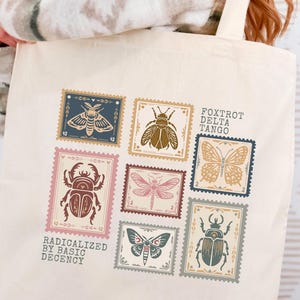 May include: Beige tote bag featuring a collection of insect-themed postage stamp designs in various colors. The stamps include images of beetles, butterflies, and dragonflies. The bag also has the text "FOXTROT DELTA TANGO" and "RADICALIZED BY BASIC DECENCY."