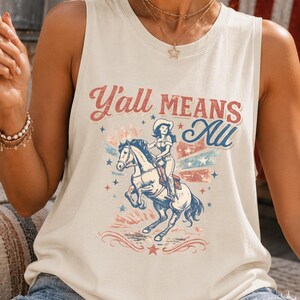 Op de afbeelding: Crèmekleurige tanktop met de tekst "Y'all Means All" en een afbeelding van een cowgirl op een paard. Het ontwerp bevat rode, witte en blauwe elementen. De persoon houdt een Amerikaanse vlag vast.