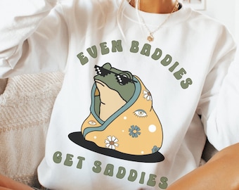 May include: White sweatshirt with a green frog wearing sunglasses and a yellow and white patterned blanket. The text on the sweatshirt reads "Even Baddies Get Saddies".