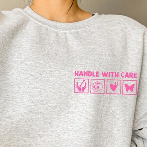 Könnte beinhalten: Hellgraues Sweatshirt mit dem pinkfarbenen Schriftzug "HANDLE WITH CARE" und pinkfarbenen grafischen Symbolen einer Hand, eines Schädels, eines Herzens und eines Schmetterlings. Das Sweatshirt ist Teil eines passenden Sets.