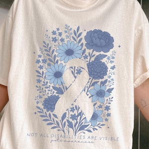 Pode incluir: Camiseta creme com um design de fita azul e elementos florais azuis. A camiseta tem o texto "NOT ALL DISABILITIES ARE VISIBLE" e "pete awareness" impresso. É um estilo casual de manga curta.