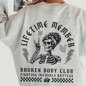 以下が含まれることがあります： 黒い蝶と「Lifetime Member Broken Body Club Fighting Invisible Battles」というテキストが書かれた、白黒のスケルトンが描かれたグレーのトレーナーです。