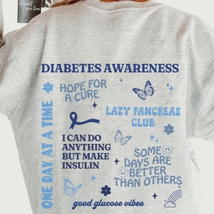 May include: Grey sweatshirt with a blue and white graphic print that says "Diabetes Awareness" and includes a blue ribbon, butterflies, and the text "One Day at a Time", "Hope for a Cure", "Lazy Pancreas Club", "I can do anything but make insulin", "Some days are better than others", and "Good glucose vibes".