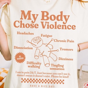 以下が含まれることがあります： 「My Body Chose Violence」というテキストと、ピンが刺さったブードゥー人形のグラフィックが描かれた白いTシャツ。グラフィックには、「頭痛、疲労、慢性的な痛み、震え、めまい、歩行困難、しびれ」というテキストも含まれています。「24時間365日痛みを感じています。あなたが見ていないからといって、存在しないわけではありません。どうか優しくしてください！」というテキストもシャツに書かれています。シャツの下部には市松模様があり、その下に「Have a nice day!」というテキストが印刷されています。