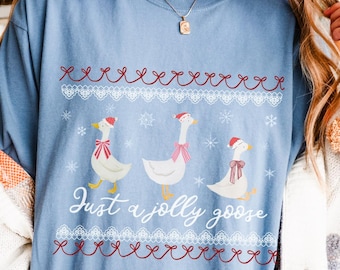 May include: A light blue t-shirt featuring a festive design with three geese wearing Santa hats and scarves. The text "Just a jolly goose" is written below the geese. The shirt has a decorative red and white pattern.