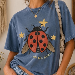 May include: A blue short-sleeve t-shirt featuring a graphic of a red ladybug with black spots, surrounded by yellow stars. The text "SAD BUG SUMMER" is printed in yellow below the ladybug. The shirt is worn with light blue denim jeans.