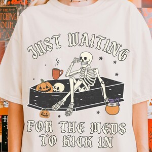 May include: A cream colored t-shirt with a skeleton in a coffin and the text "Just Waiting for the meds to kick in".