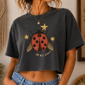 May include: A charcoal gray cropped t-shirt featuring a graphic of a red ladybug with black spots, surrounded by three yellow stars. The text "SAD BUG SUMMER" is printed in yellow below the ladybug. The shirt has short sleeves and a crew neckline.