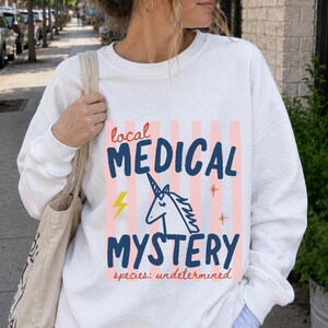 Op de afbeelding: Een wit sweatshirt met lange mouwen en een afbeelding met verticale roze strepen, een blauwe eenhoornillustratie en de tekst "local MEDICAL MYSTERY species: undetermined" in blauw en rood, vergezeld van een gele bliksemschicht en rode sterren.