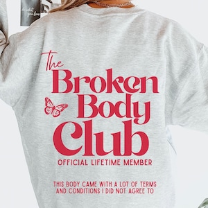 Può includere: Una felpa grigio chiaro con il testo rosso "The Broken Body Club" e una grafica a farfalla. La felpa include anche le frasi "OFFICIAL LIFETIME MEMBER" e "THIS BODY CAME WITH A LOT OF TERMS AND CONDITIONS I DID NOT AGREE TO."