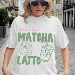 May include: White t-shirt with the text "fueled by MATCHA LATTE" in green and pink. The design includes illustrations of matcha preparation and an iced drink. A beige cap with "The Western House Casino" is also visible.