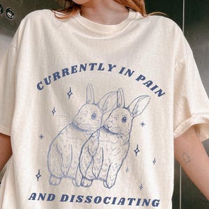 Może przedstawiać: Kremowy t-shirt z niebieską ilustracją dwóch królików i napisem "CURRENTLY IN PAIN AND DISSOCIATING". Koszulka ma krótkie rękawy i luźny krój.