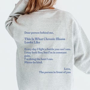 Puede incluir: Sudadera gris ceniza con el texto: "Querida persona detrás de mí, así es como se ve la enfermedad crónica". El texto continúa con un mensaje sobre la lucha diaria. La sudadera se combina con una gorra naranja.