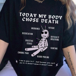 May include: Black t-shirt featuring a graphic of a voodoo doll with pins in it. The text "TODAY MY BODY CHOSE DEATH" is above the doll. The text "I am in pain 24/7. Just because you can't see it, it doesn't mean it doesn't exist." is below the doll.