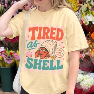 Könnte beinhalten: Buttergelbes T-Shirt mit dem Text "TIRED as SHELL" in Orange, Rosa und Türkis. Die Grafik zeigt einen Cartoon-Einsiedlerkrebs in einer Muschel, eine Muschel und einen Seestern. Das Shirt hat kurze Ärmel.