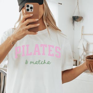 May include: White t-shirt with the text "PILATES & matcha" in pink and green lettering. The model is wearing a black cap and holding a brown bowl. The shirt has a relaxed, oversized fit.