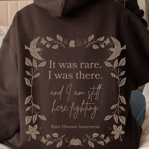May include: A brown hooded sweatshirt with a floral design and the text "It was rare. I was there. and I am still here fighting. Rare Disease Awareness."