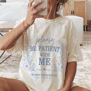 May include: A cream-coloured t-shirt featuring the light blue text "please BE PATIENT WITH ME" within a floral wreath. The shirt also has the text "MY BODY FIGHTS BATTLES you cannot see". The person is wearing white socks with black stripes.