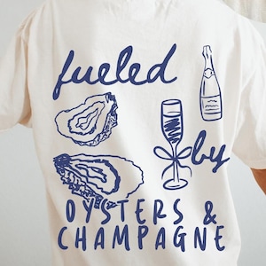 Puede incluir: Camiseta blanca con un gr&aacute;fico azul que dice "fueled by oysters & champagne" con ilustraciones de ostras, una botella de champ&aacute;n y una copa de champ&aacute;n.